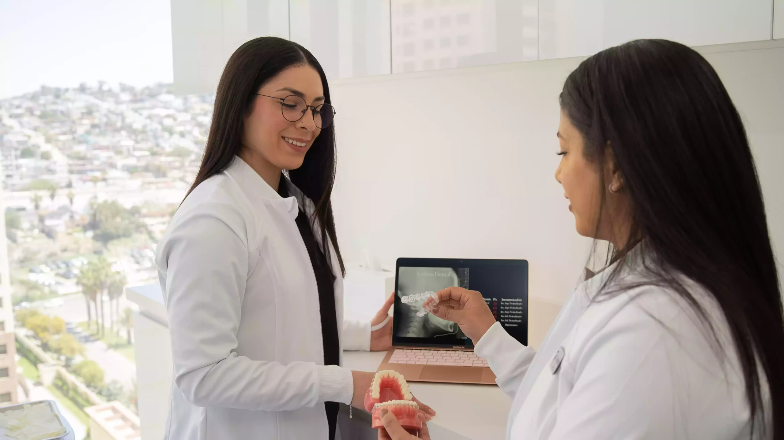 Two women in white lab coats discuss dental care, one holding a dental model and the other pointing at a dental X-ray on a laptop.