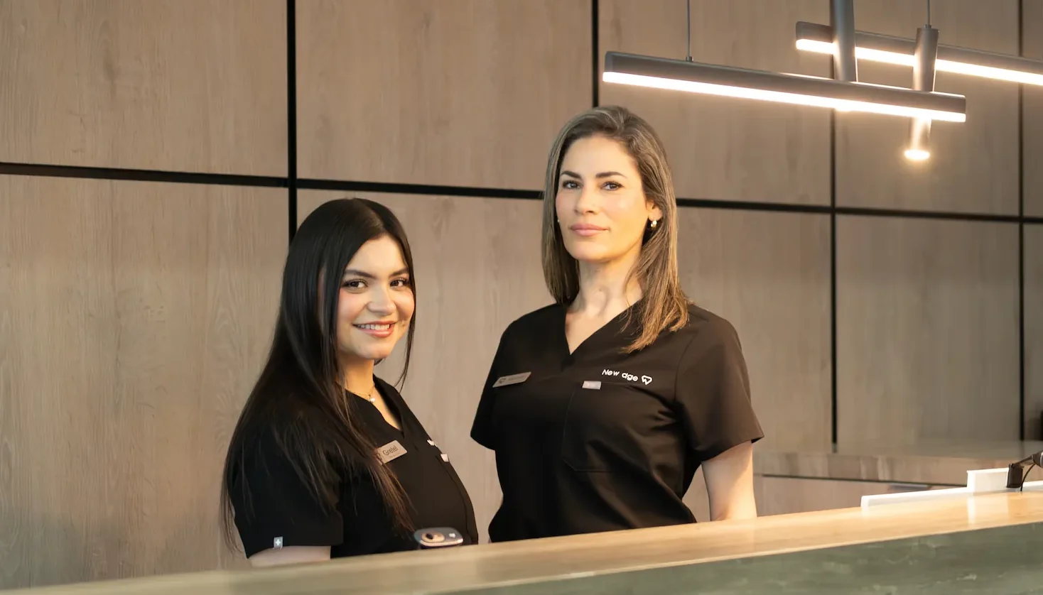 Two women in black uniforms stand behind a marble reception desk in a modern, wood-paneled office space.