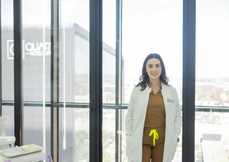 A woman in a white lab coat stands in front of large windows in a modern office or medical setting, smiling at the camera.