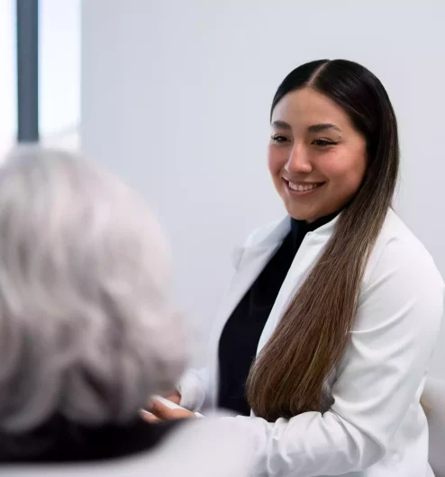 A young woman in a white coat smiles while speaking with an older person with gray hair in an office setting.