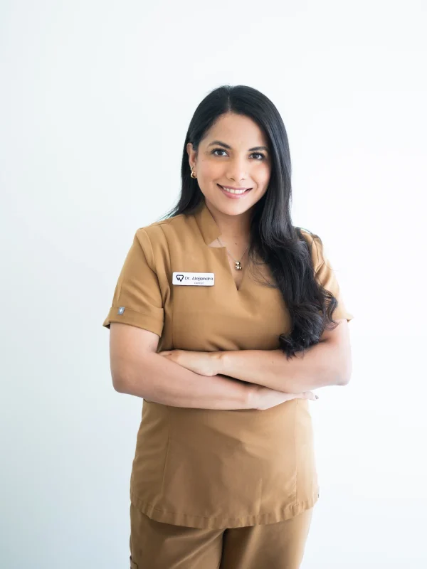 A woman wearing brown medical scrubs with a name tag stands with arms crossed, smiling in front of a plain white background.