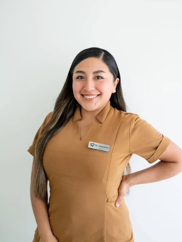 A woman with long brown hair, wearing a brown uniform and a name tag, stands smiling with one hand on her hip in front of a plain white background.