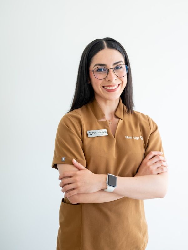 A woman with long dark hair, glasses, and a brown uniform stands against a white background, smiling with arms crossed and wearing a smartwatch.