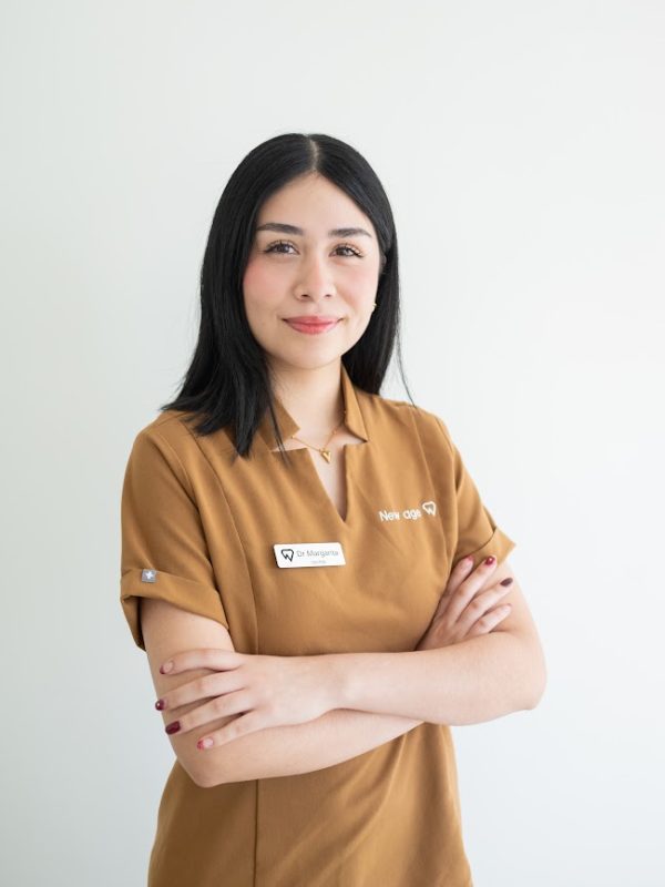 A woman with straight black hair stands against a white background, wearing a brown uniform with a name tag and her arms crossed, smiling slightly.