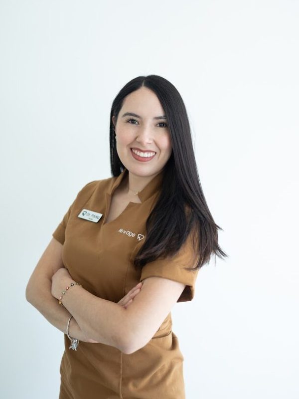 A woman with long dark hair, wearing a brown uniform and a name tag, stands against a plain white background with her arms crossed and smiling at the camera.