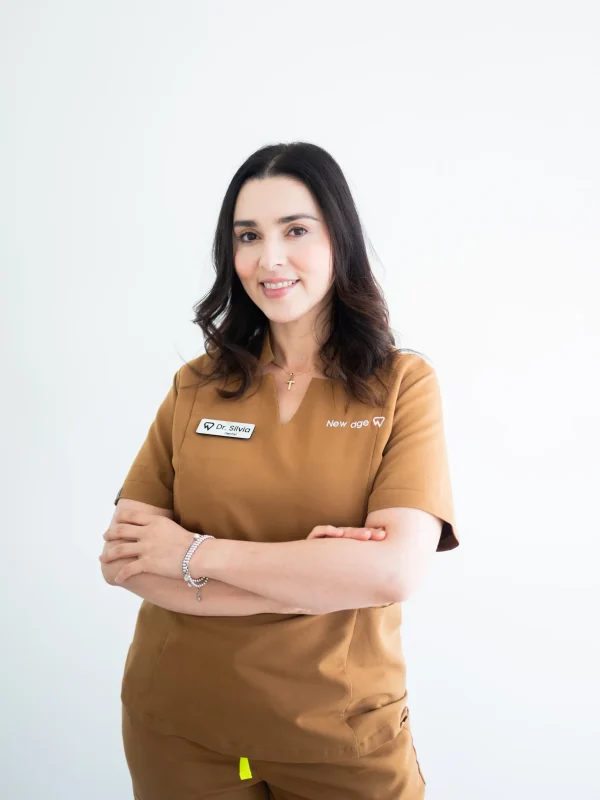 A woman in brown scrubs with name tags stands against a plain white background, smiling with her arms crossed.