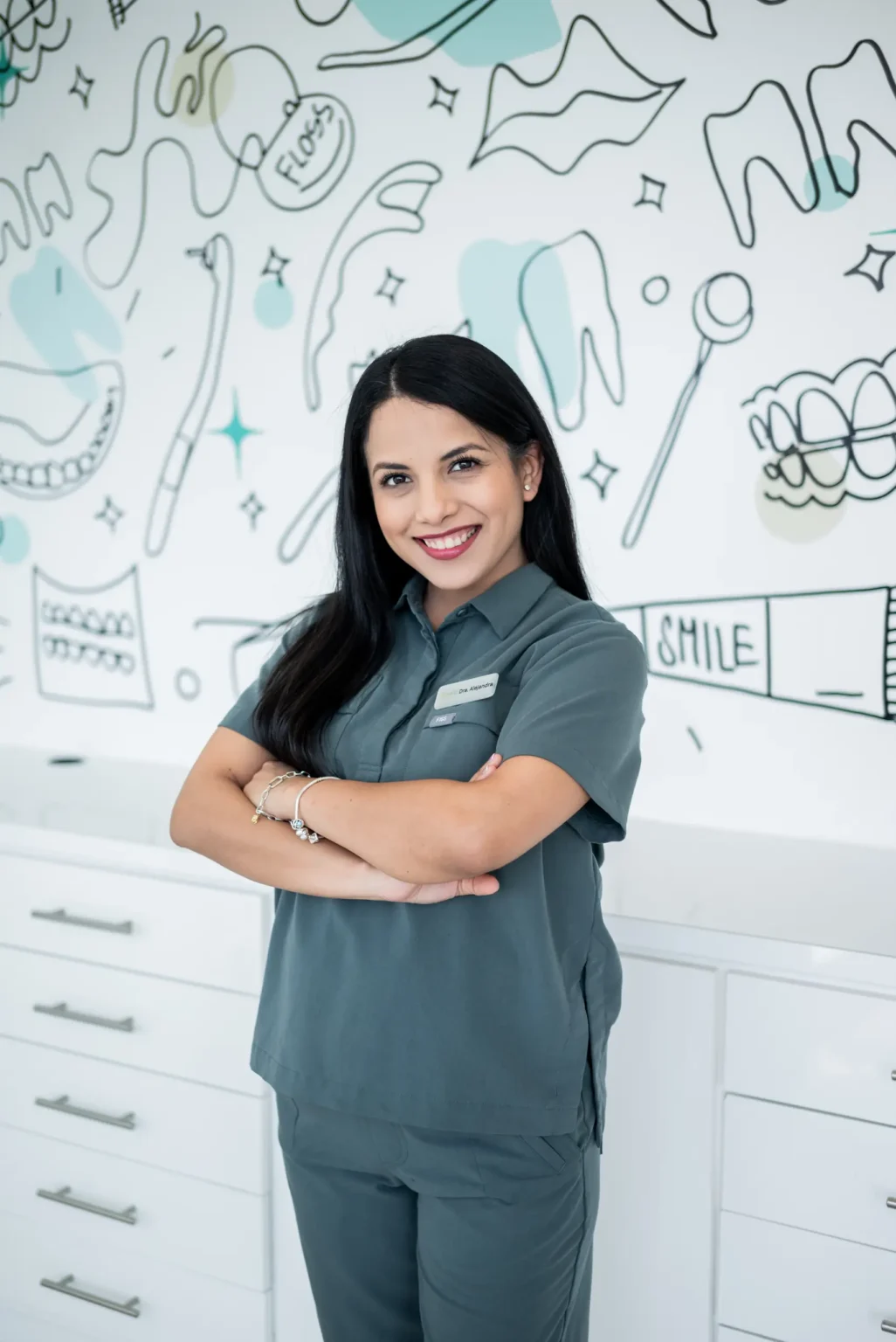 A woman in dental scrubs smiling confidently in front of a mural with dental-themed illustrations.