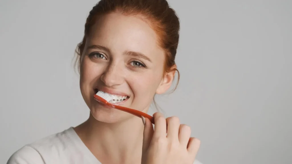 Young woman with red hair brushing her teeth, smiling at the camera, holding an orange toothbrush; light gray background. She uses baking soda to whiten her teeth.
