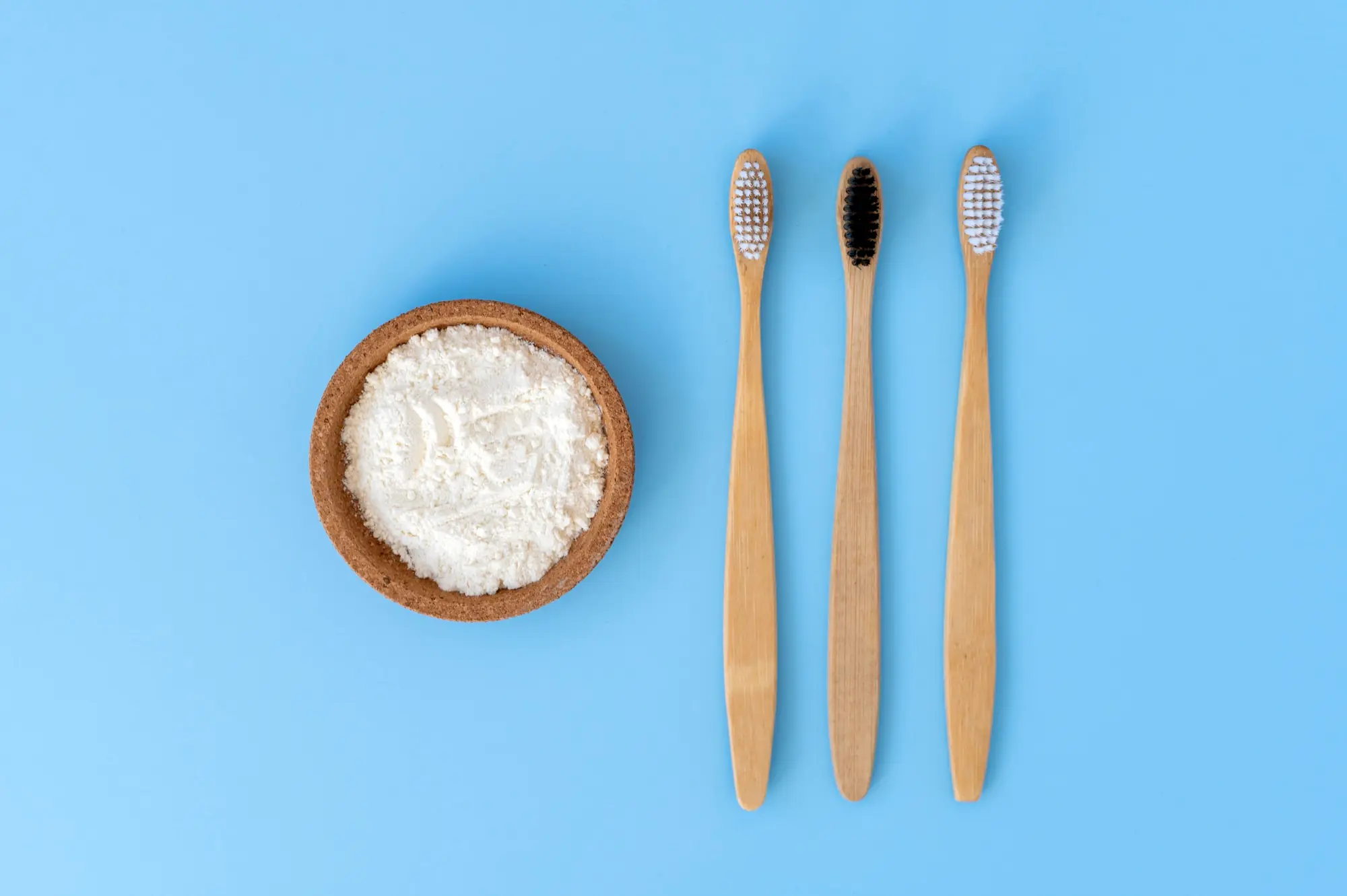 Two bamboo toothbrushes next to a small bowl of baking soda on a blue background.