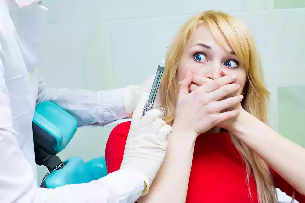 Understanding Dental Anxiety: Overcoming the Fear of the Dentist 36 A dentist prepares to administer an injection while a patient with blonde hair covers her mouth with her hands, looking anxious, as if drafting the courage for what's to come.