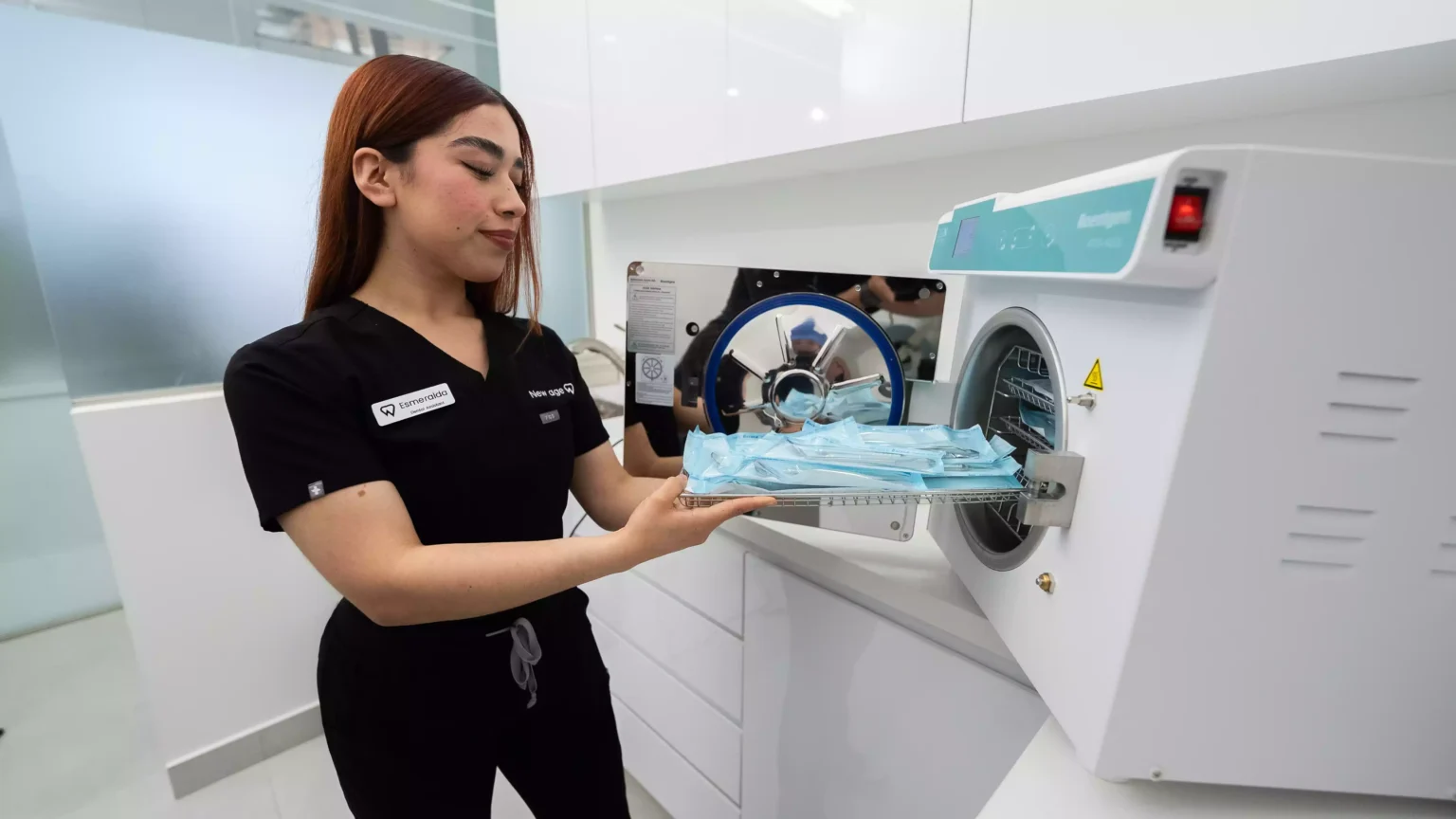 A woman in scrubs is placing a tray of bagged medical instruments into an autoclave sterilizer in a clinical setting.