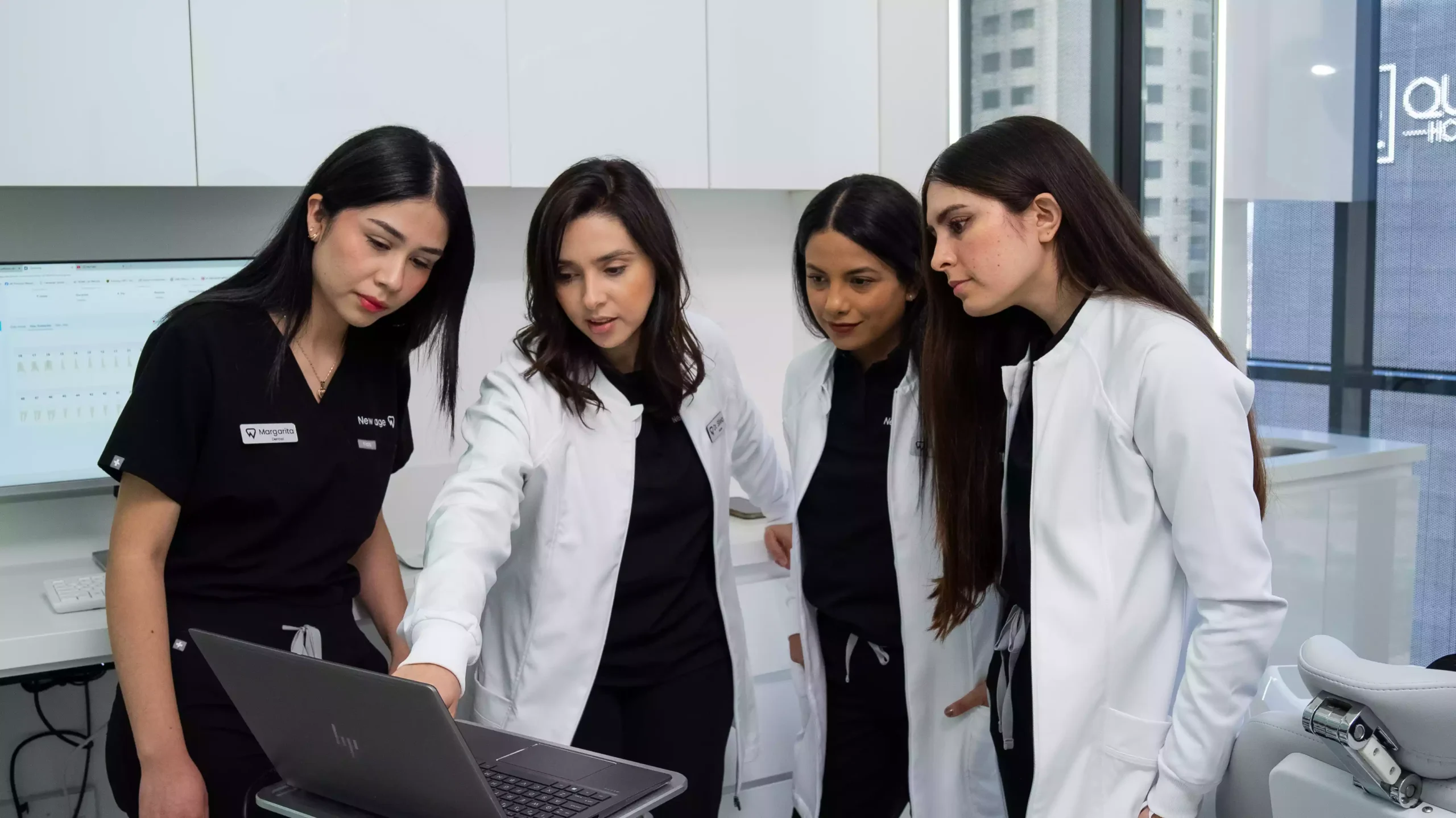 Four women in medical attire stand in a modern clinic, closely looking at a laptop screen, with medical equipment and large windows in the background.