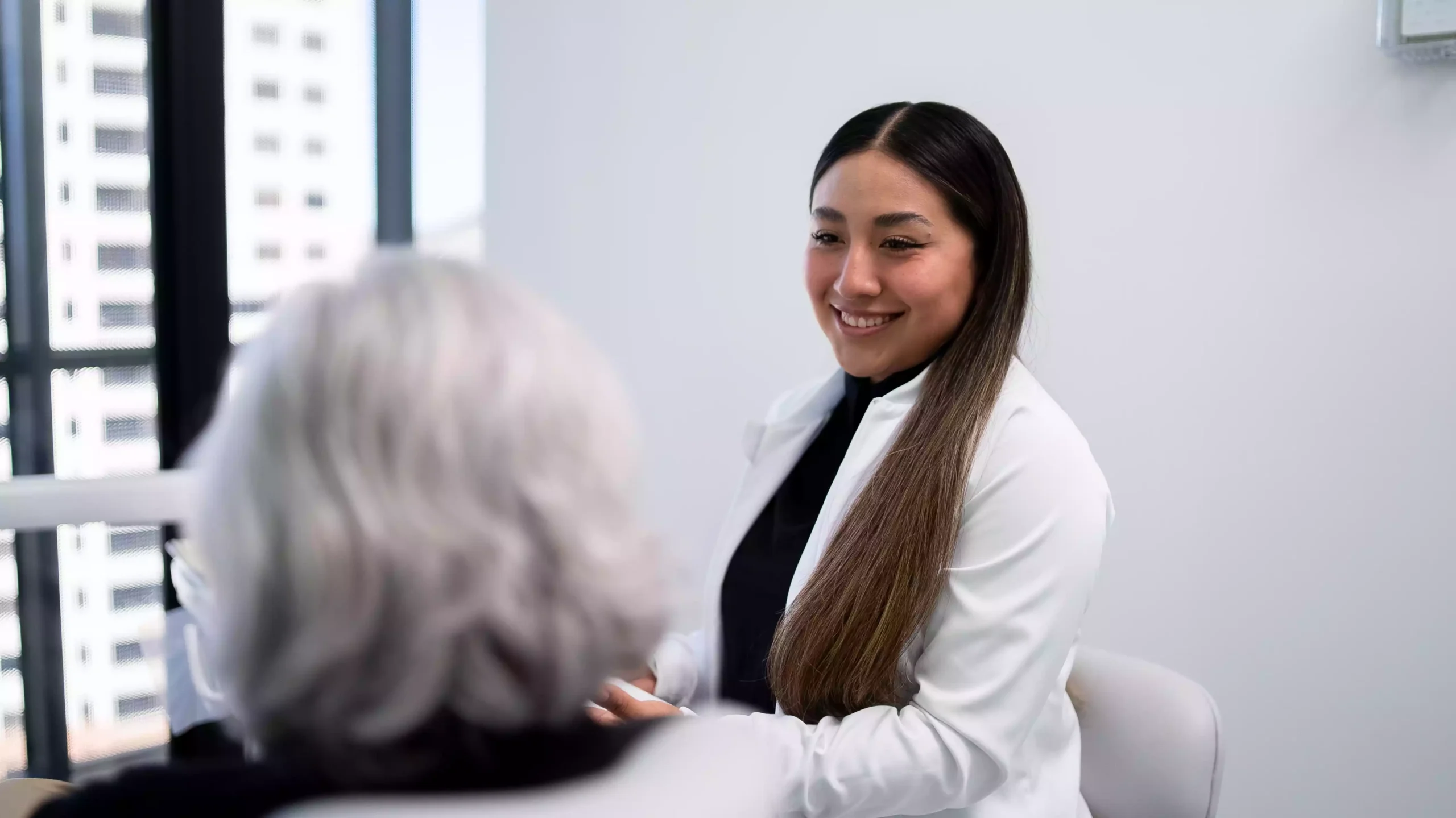 A young woman in a white coat smiles while speaking with an older person with gray hair in an office setting.