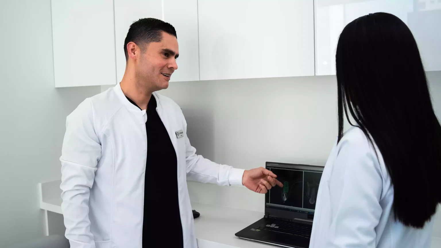 Two people in white lab coats stand near a laptop displaying medical images, having a discussion in a clinical setting.