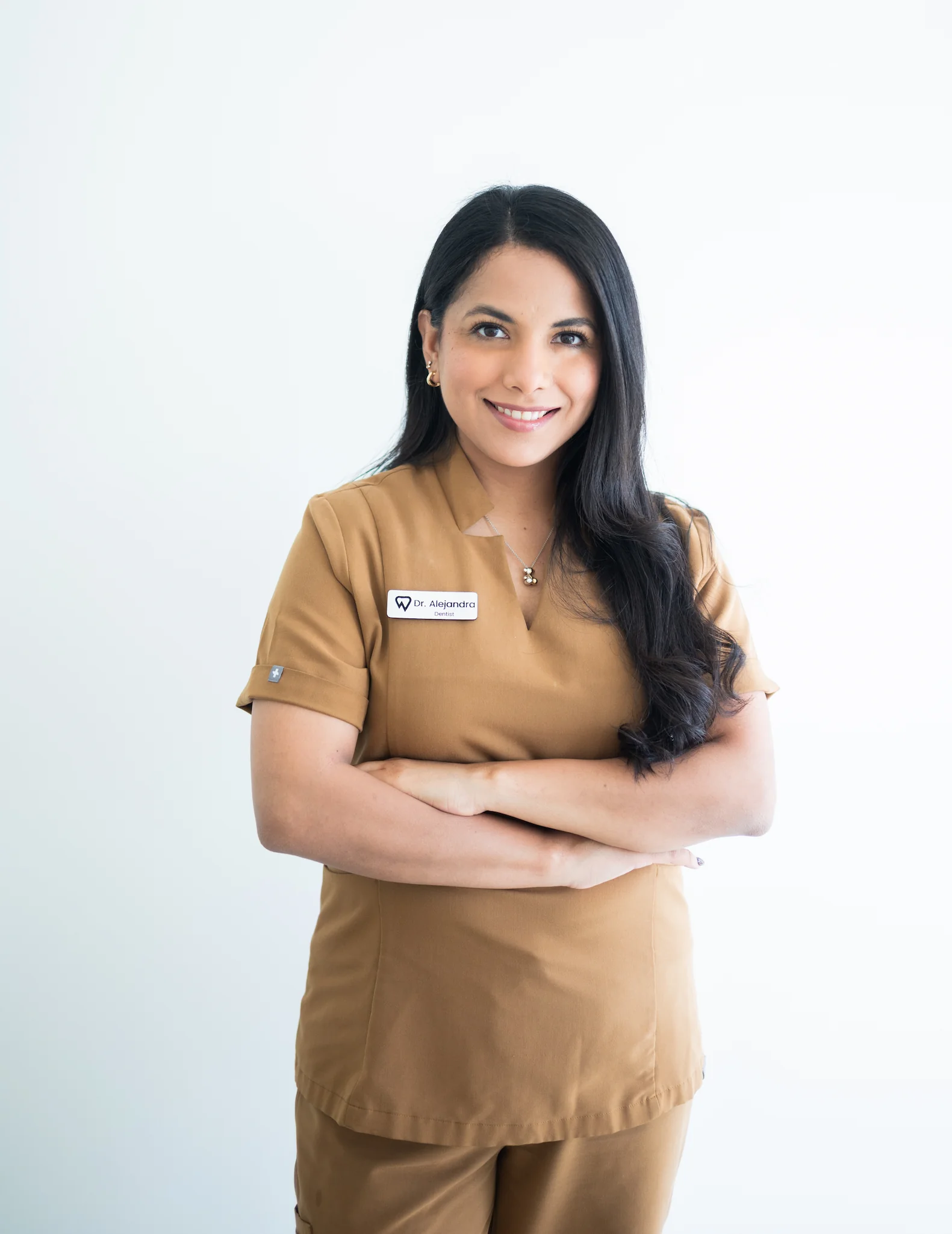 A woman wearing brown medical scrubs with a name tag stands with arms crossed, smiling in front of a plain white background.
