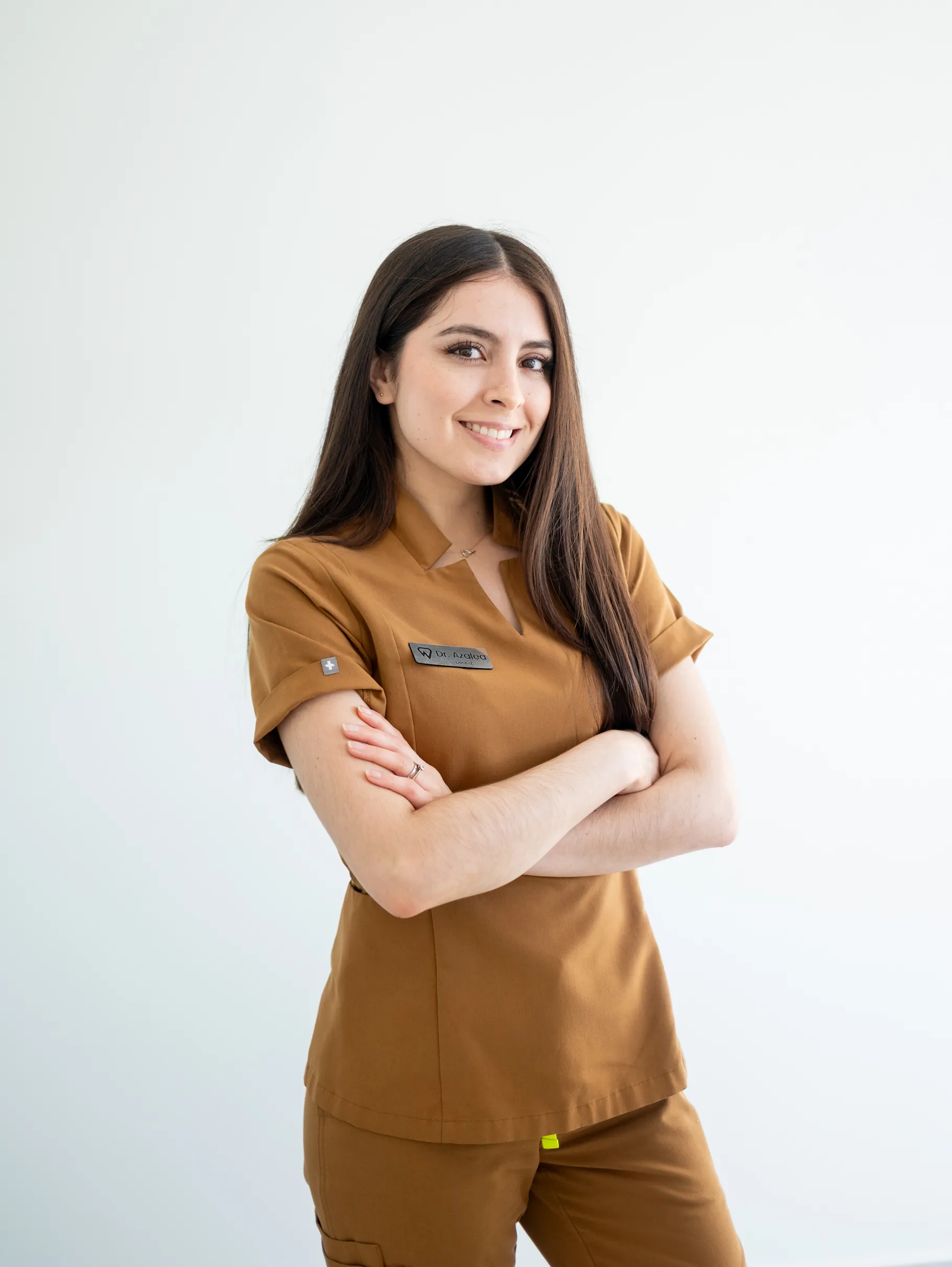 A woman with long dark hair, wearing brown medical scrubs, stands with arms crossed and smiles at the camera against a plain white background.