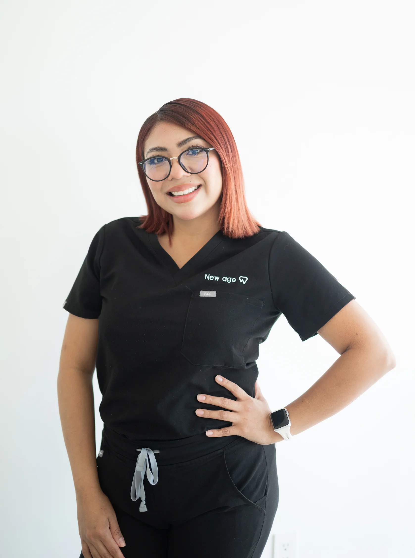 A person wearing glasses and black scrubs stands against a white background, smiling with one hand on their hip.