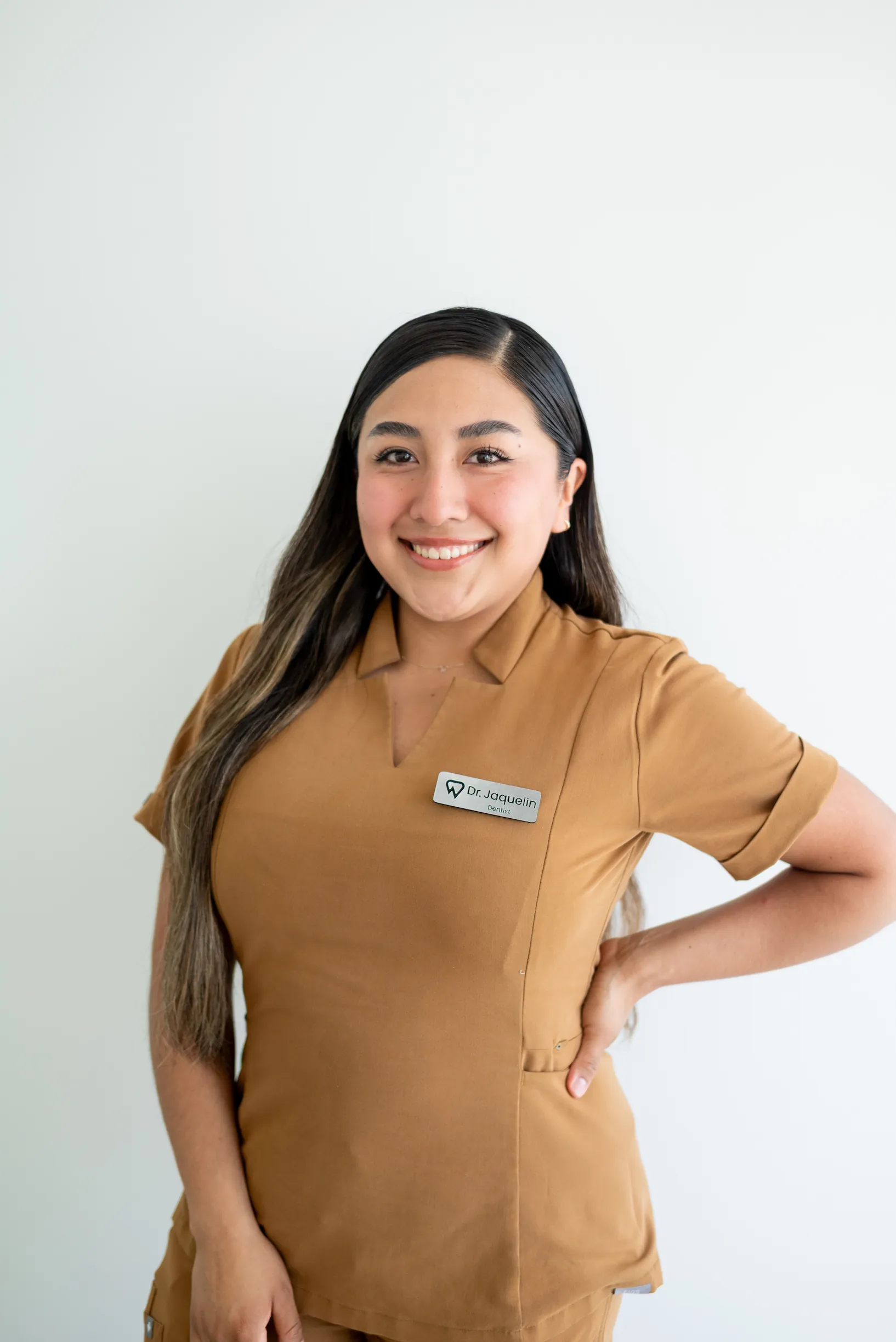 A woman with long brown hair, wearing a brown uniform and a name tag, stands smiling with one hand on her hip in front of a plain white background.