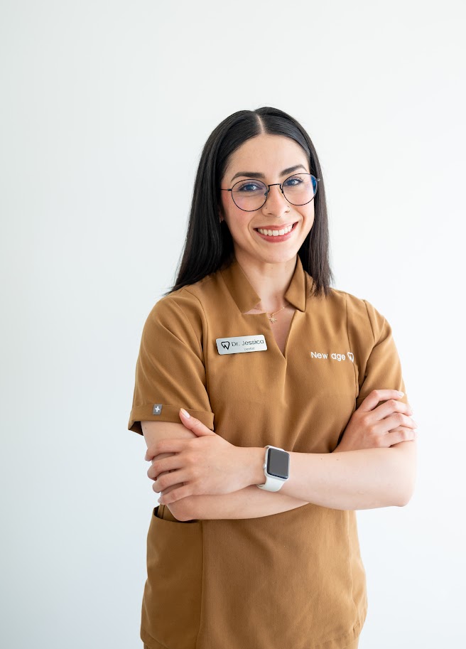 A woman with long dark hair, glasses, and a brown uniform stands against a white background, smiling with arms crossed and wearing a smartwatch.