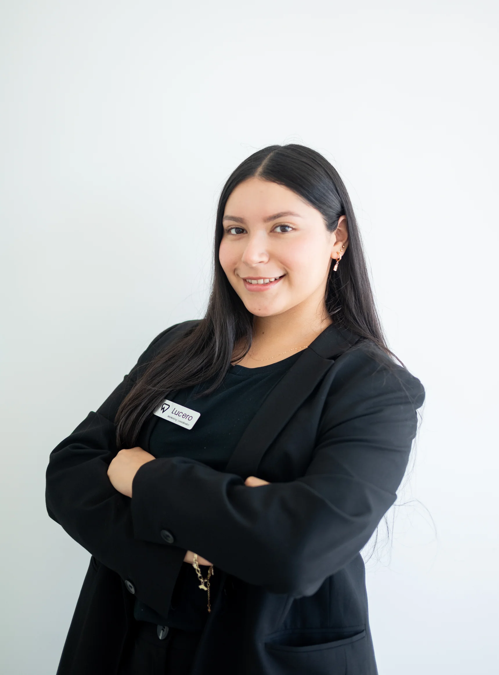 Woman with long dark hair wearing a black blazer stands with arms crossed, smiling at the camera against a plain white background. She has a name tag on her blazer.