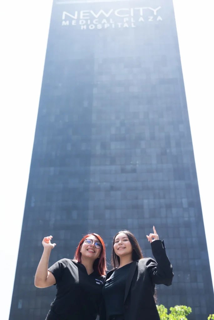 Two women stand in front of a tall, dark glass building labeled "New City Medical Plaza Hospital," smiling and gesturing upward.