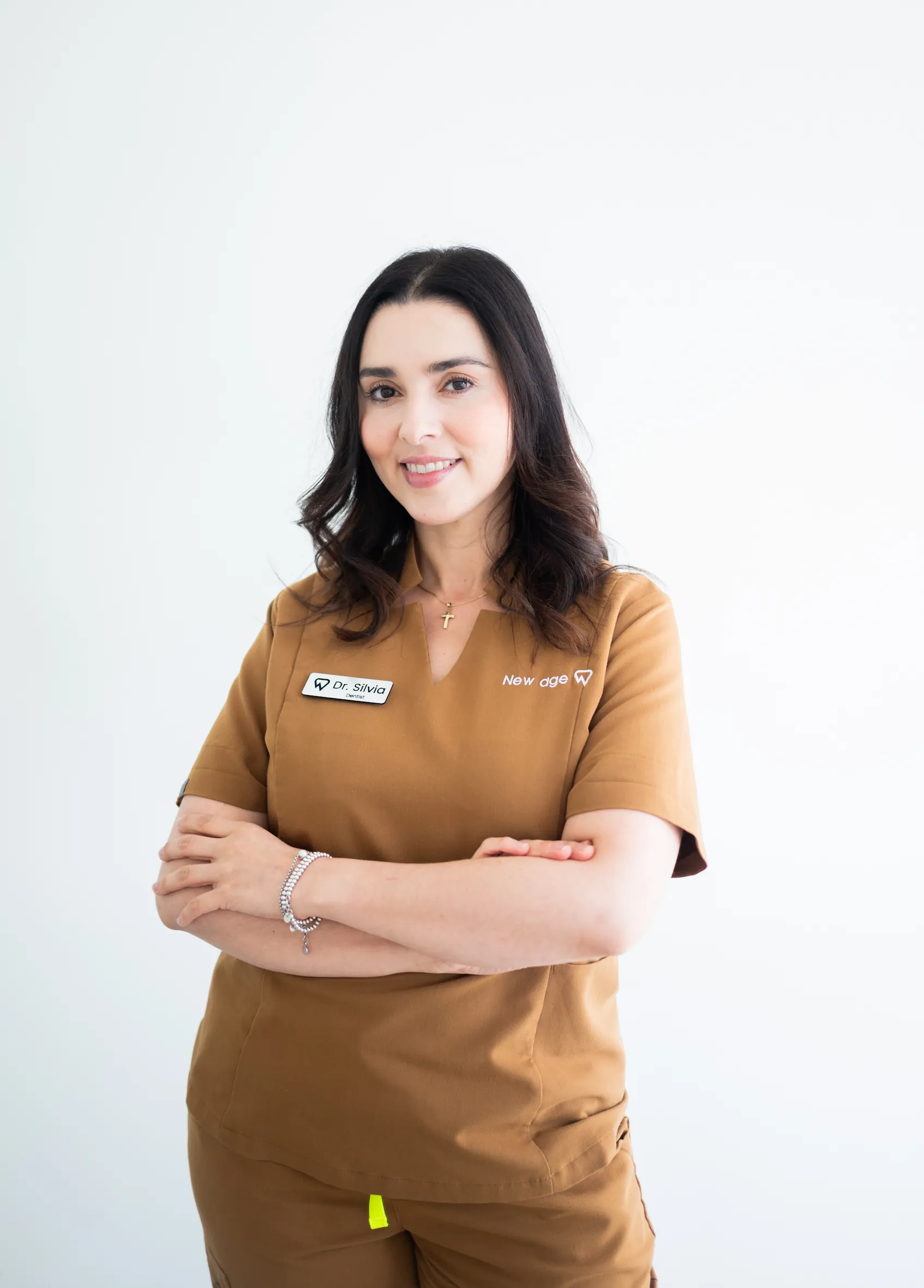 A woman in brown scrubs with name tags stands against a plain white background, smiling with her arms crossed.