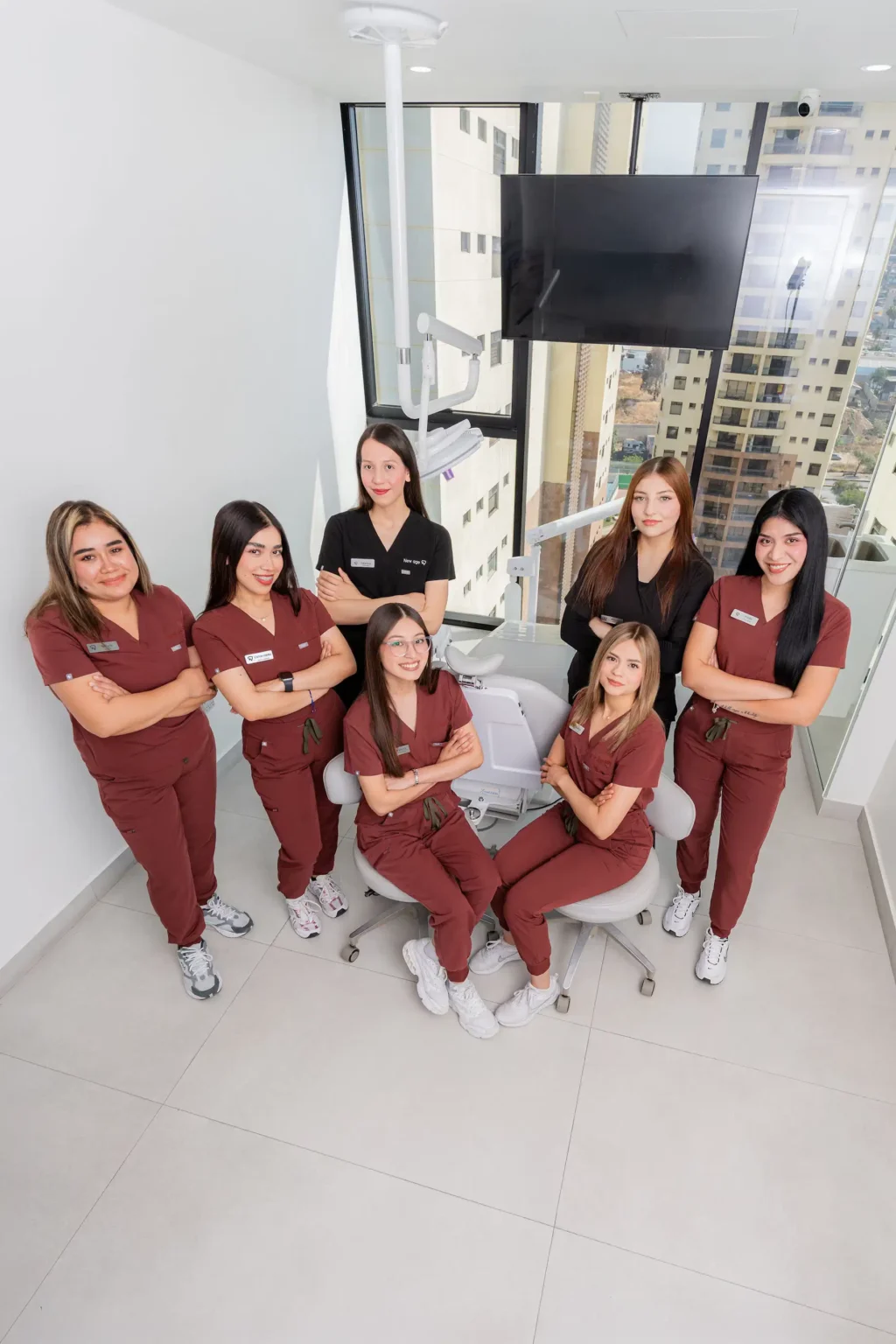 Seven dental professionals in uniforms pose together in a modern dental office, standing and sitting around a dental chair with large windows in the background.