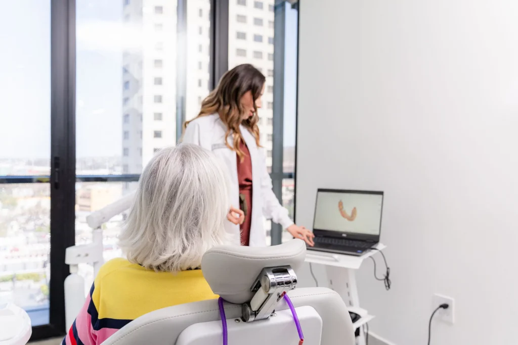 A dentist consults with a patient, showing an all on 4 dental image on a laptop in a bright office with large windows and city views.