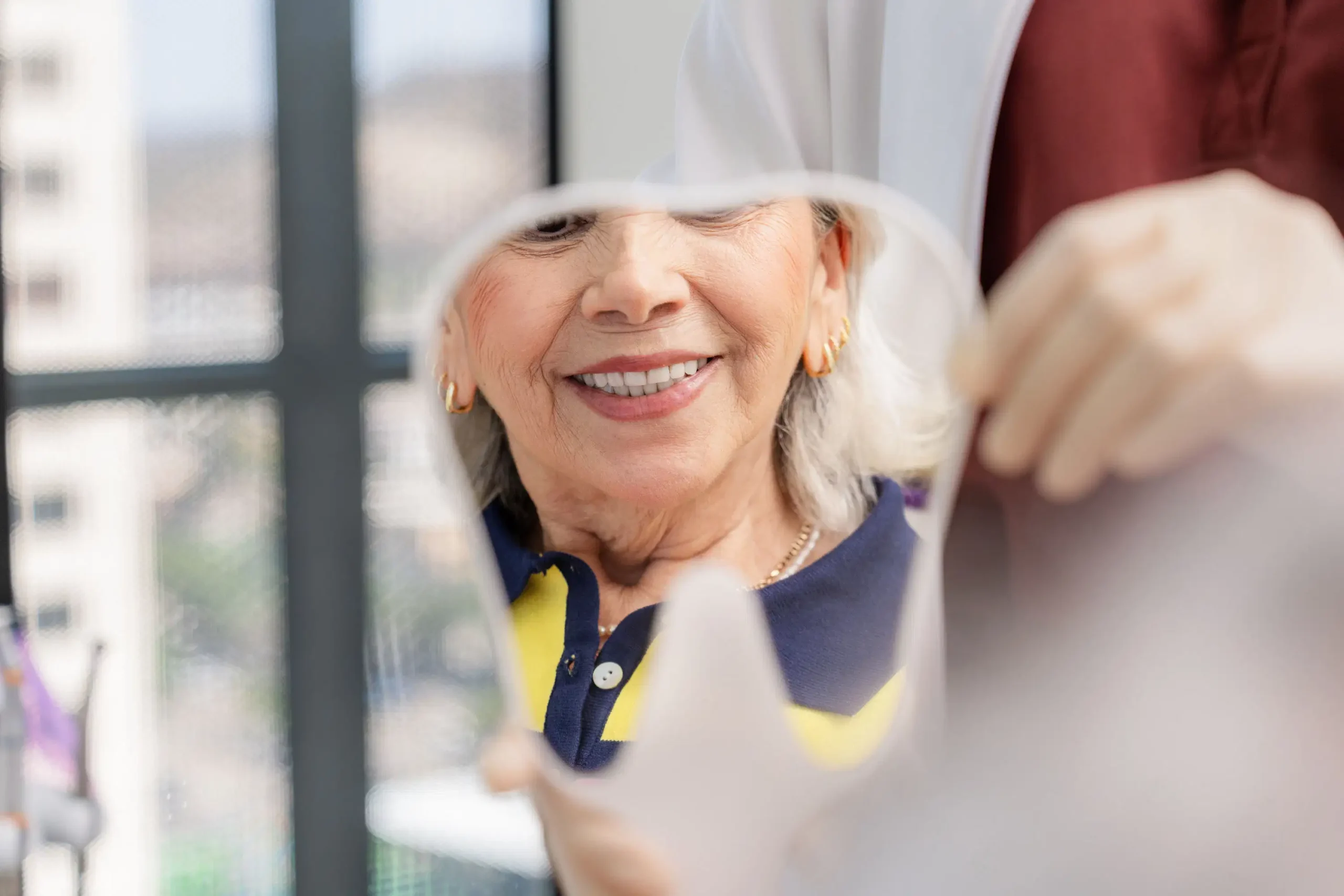 An older woman smiles as she admires her reflection in a dental mirror held by a gloved person, appreciating the results of Smile Design, with city buildings visible through the windows in the background.