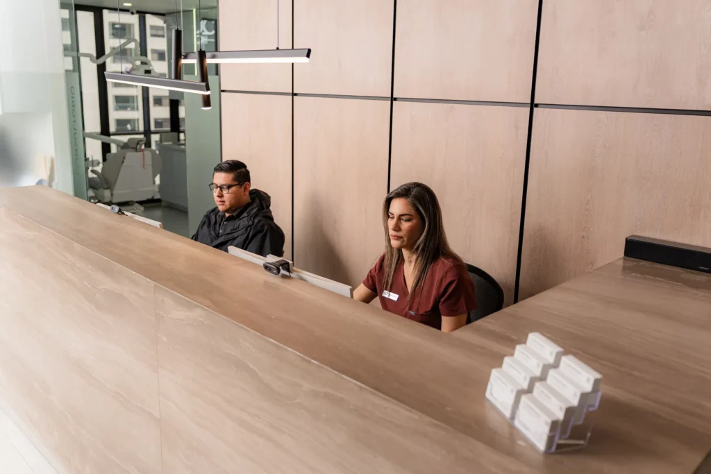 Two receptionists sit behind a modern, wooden reception desk, working on computers in a brightly lit office environment.