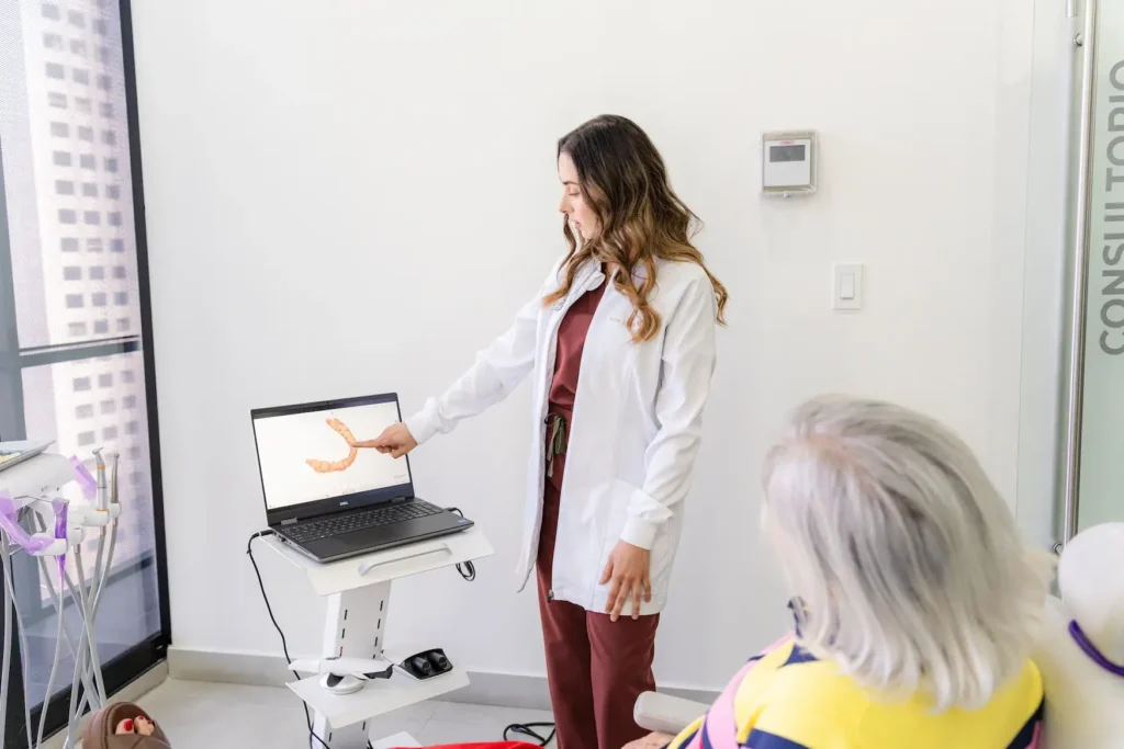 A doctor in a lab coat points to a digestive tract diagram on a laptop screen while speaking to a seated patient in a medical office.