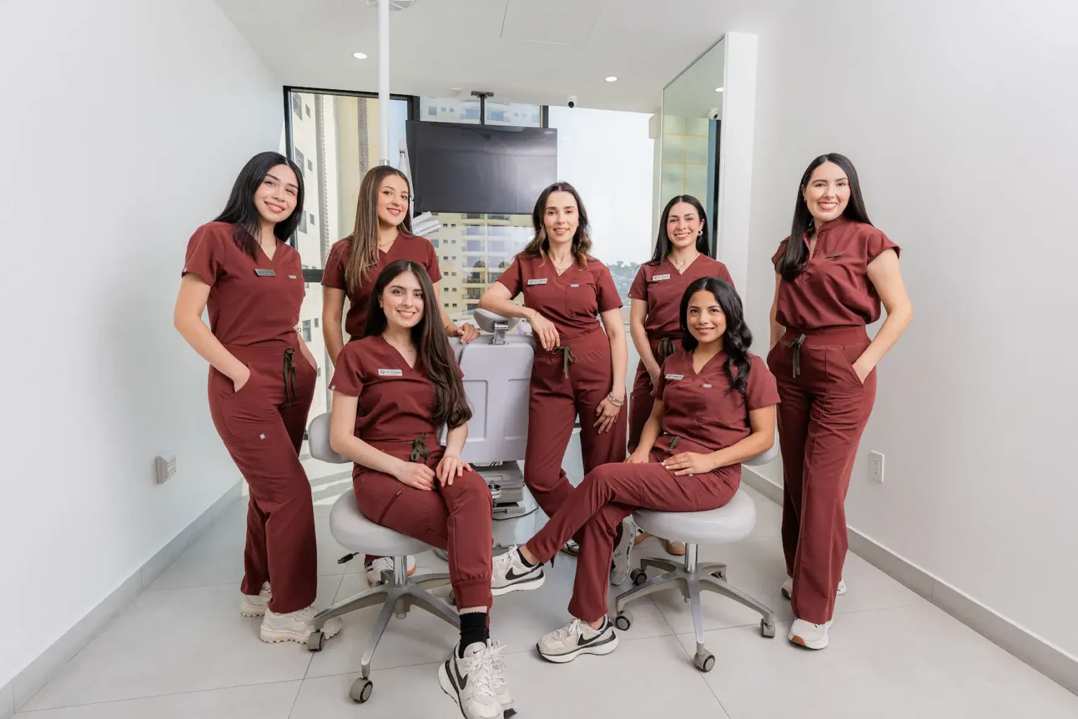 Seven women wearing matching maroon scrubs pose together in a clean, modern medical or dental office. Some are standing, while others are seated on stools.