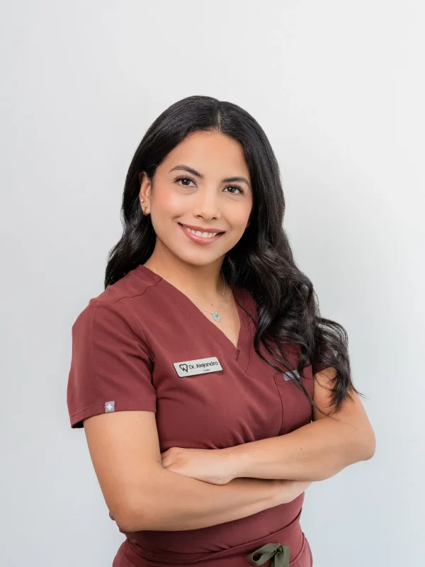 Woman in maroon medical scrubs with a name tag stands with arms crossed, smiling in front of a plain light background.