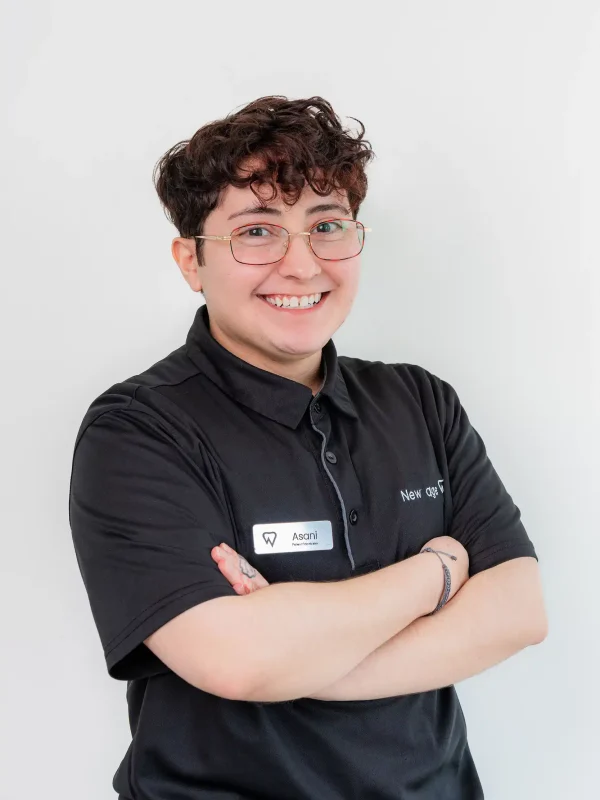 Person with short curly hair, glasses, and a name tag, wearing a black polo shirt, standing with arms crossed and smiling in front of a plain white background.
