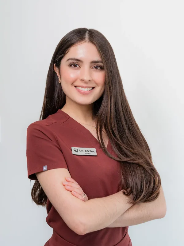 A woman with long brown hair wearing maroon scrubs and a name tag that reads "Dr. Azadad" stands with arms crossed, smiling against a plain white background.