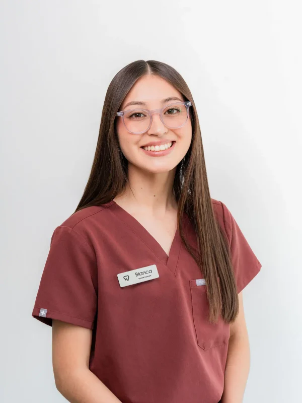 A woman with long straight hair, wearing glasses and maroon scrubs, smiles at the camera. She has a name tag on her top.