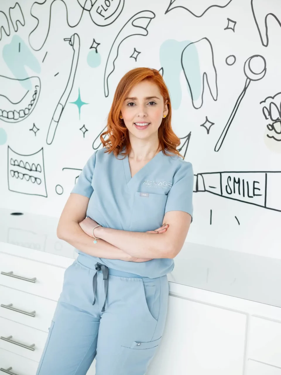 A woman in blue scrubs standing in front of a wall with doodles on it, representing a dental professional specializing in oral surgery.