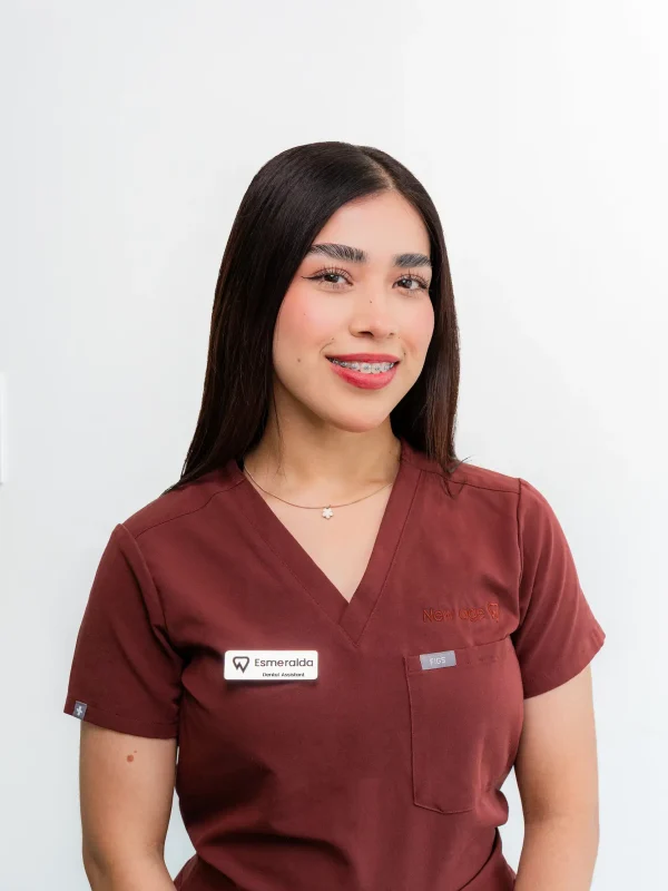 A woman with long dark hair wearing maroon scrubs and a name badge smiles at the camera against a plain white background.