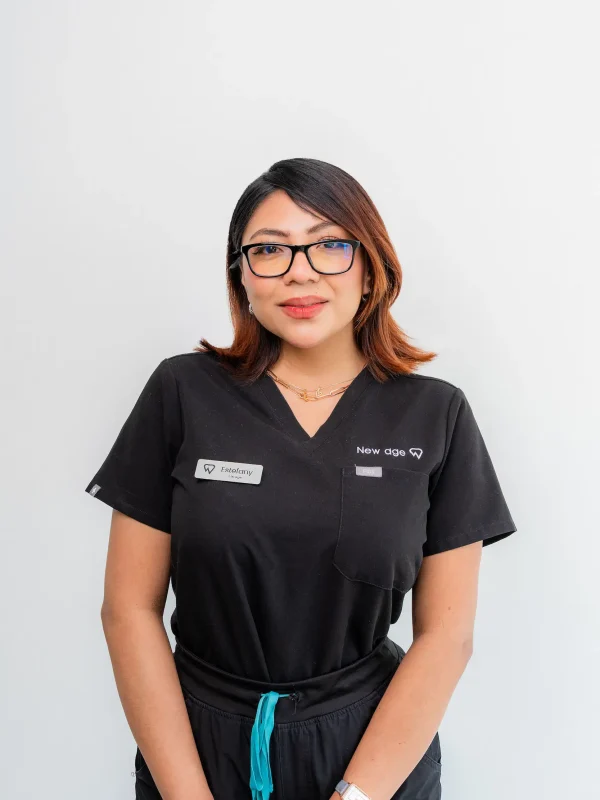 A woman with shoulder-length brown hair and glasses stands against a plain white background, wearing black scrubs with name tags that read “Fabiola V” and “New Age.”.