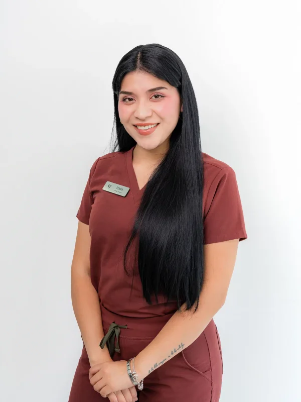 A woman with long black hair wearing maroon scrubs and a name tag stands against a plain white background, smiling at the camera.