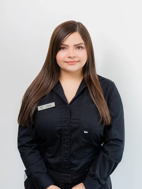 A woman with long brown hair wearing a black button-up shirt and a name tag stands against a plain white background.