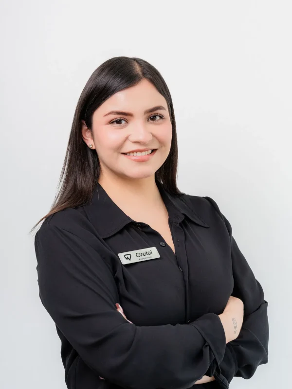 Woman with long dark hair, wearing a black collared shirt and a name tag that reads "Gretel," standing against a plain white background with arms crossed and smiling.
