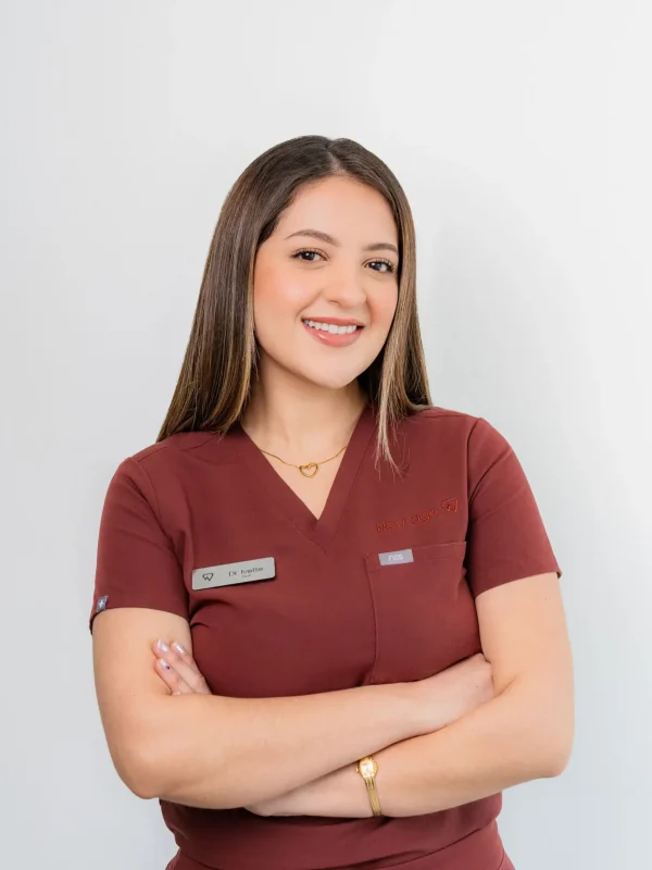Woman in maroon medical scrubs stands with arms crossed, smiling at the camera against a plain white background. She wears a name tag and gold jewelry.