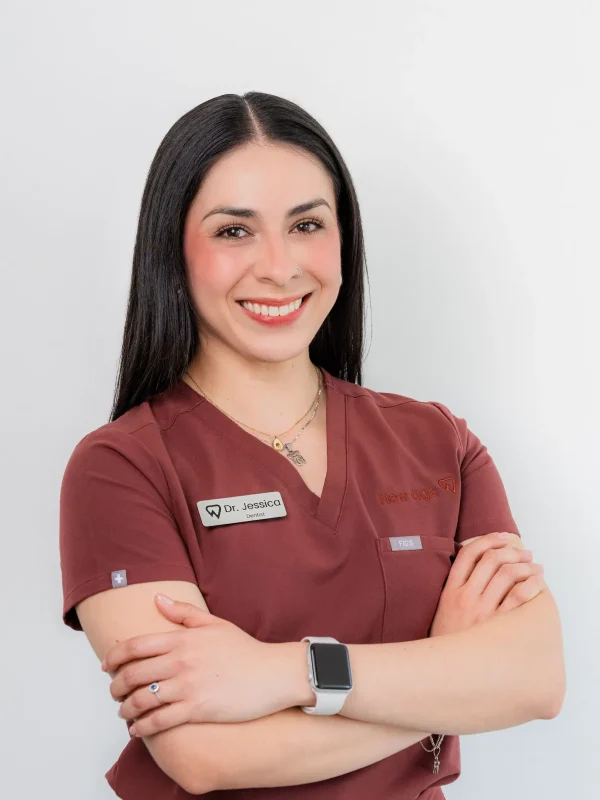 A woman wearing maroon scrubs and a name badge stands with arms crossed, smiling at the camera against a plain white background.