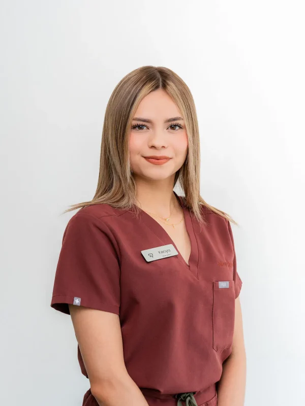 A woman wearing burgundy medical scrubs and a name tag stands against a plain white background, facing the camera with a neutral expression.