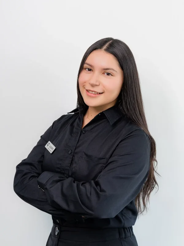 A woman with long dark hair, wearing a black button-up shirt and nametag, stands against a plain white background with arms crossed and a slight smile.
