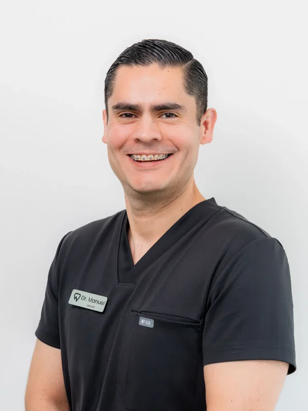 A man with short dark hair, wearing black scrubs and a name tag that says "Dr. Manuel," smiles while standing against a plain white background.