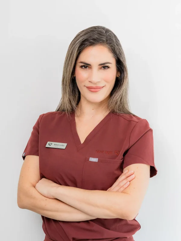 A woman in maroon medical scrubs with a name tag stands against a plain white background, arms crossed and smiling slightly.
