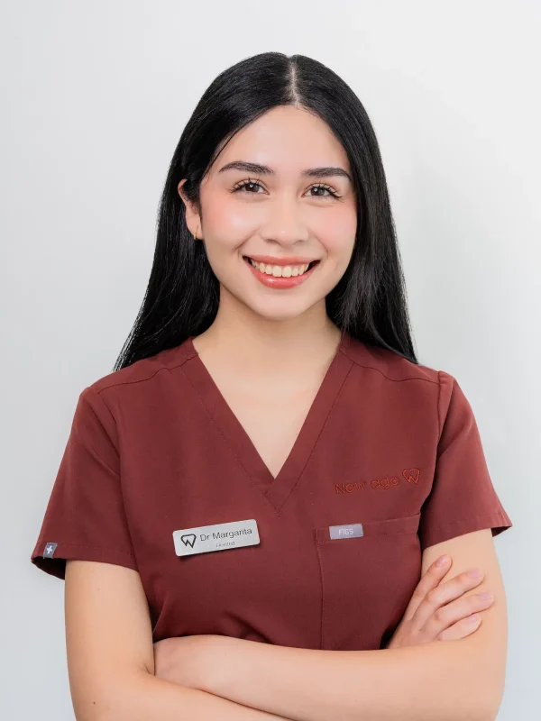 A woman wearing a maroon scrub top, name tag, and embroidered logo stands smiling with arms crossed against a plain light background.