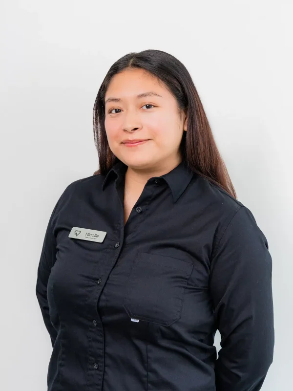 A woman with long dark hair wearing a black button-up shirt and a name tag stands against a plain white background, looking at the camera.
